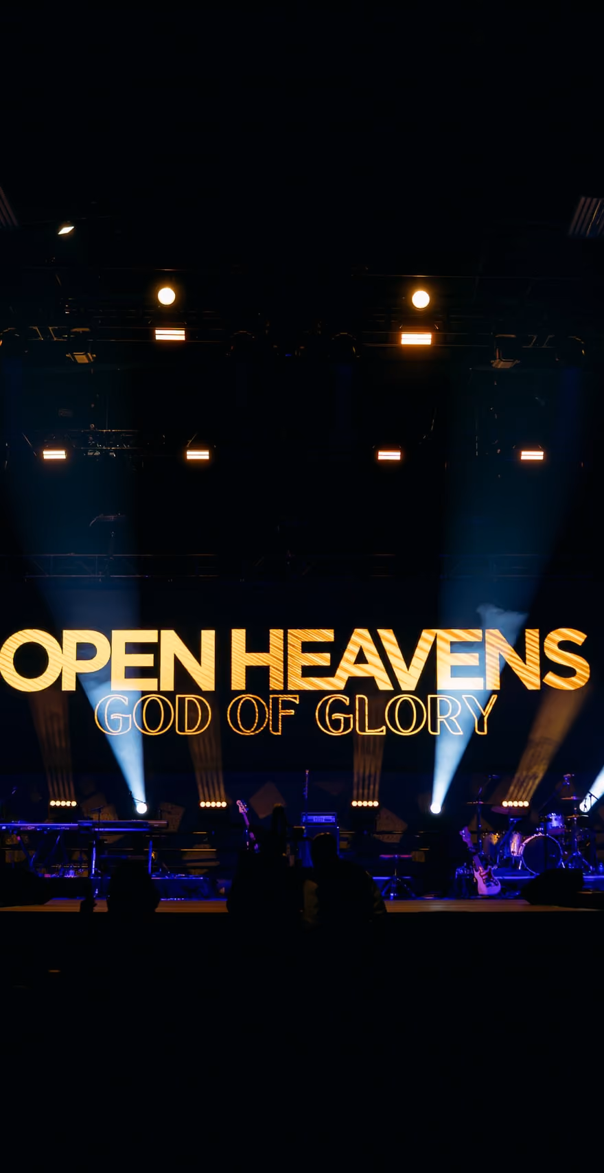 The Open Heavens stage at a previous gathering, lit in blue and gold, with the words God of Glory on the screen behind the band.