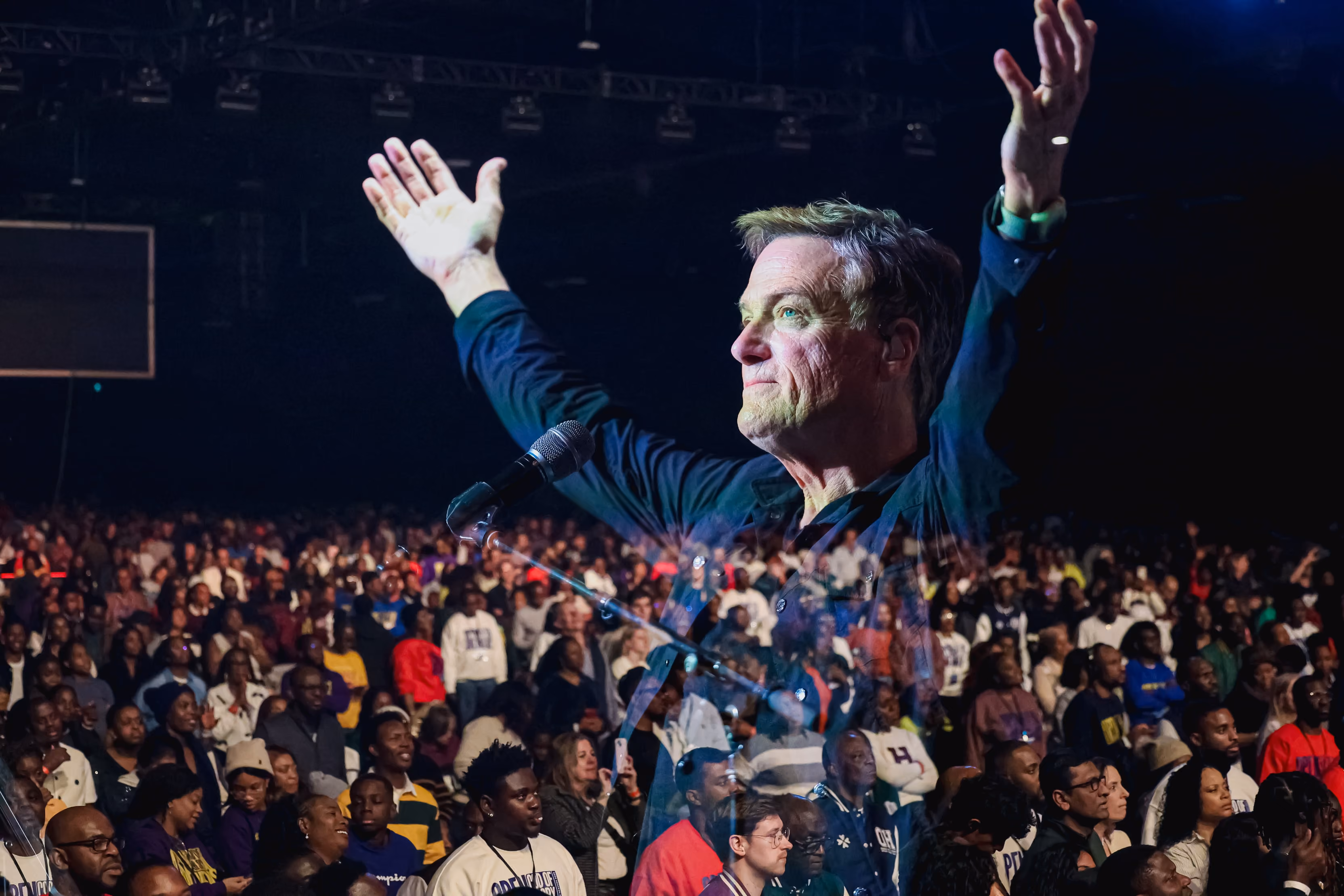 Don Moen leading worship with both hands raised, the arena crowd behind him.